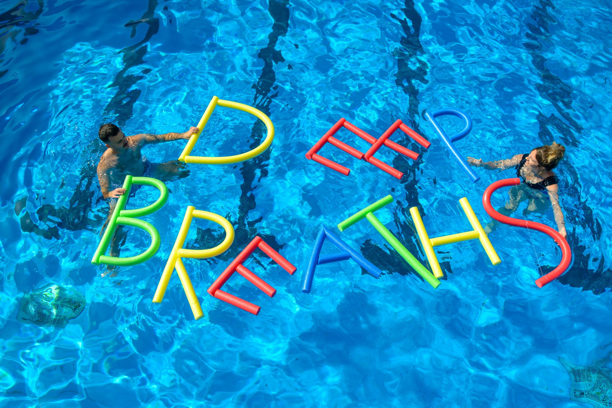 Two artists in a pool with DEEP BREATHS spelled out in the water with pool noodles.