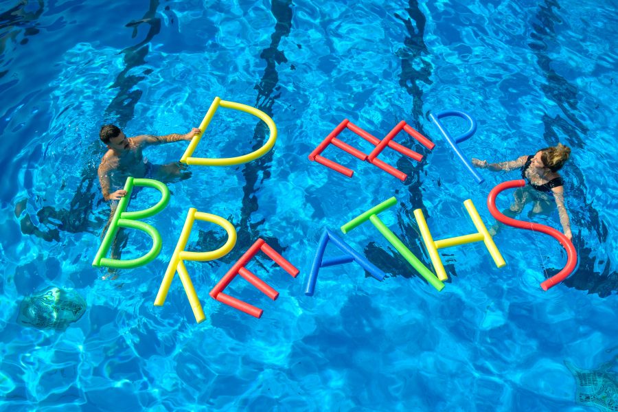 Two artists in a pool with DEEP BREATHS spelled out in the water with pool noodles.