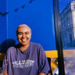 Eugenia is smiling and facing the camera. They are a mixed race person wearing a oversized casual t-shirt and they have short blonde hair. They look friendly and relaxed.