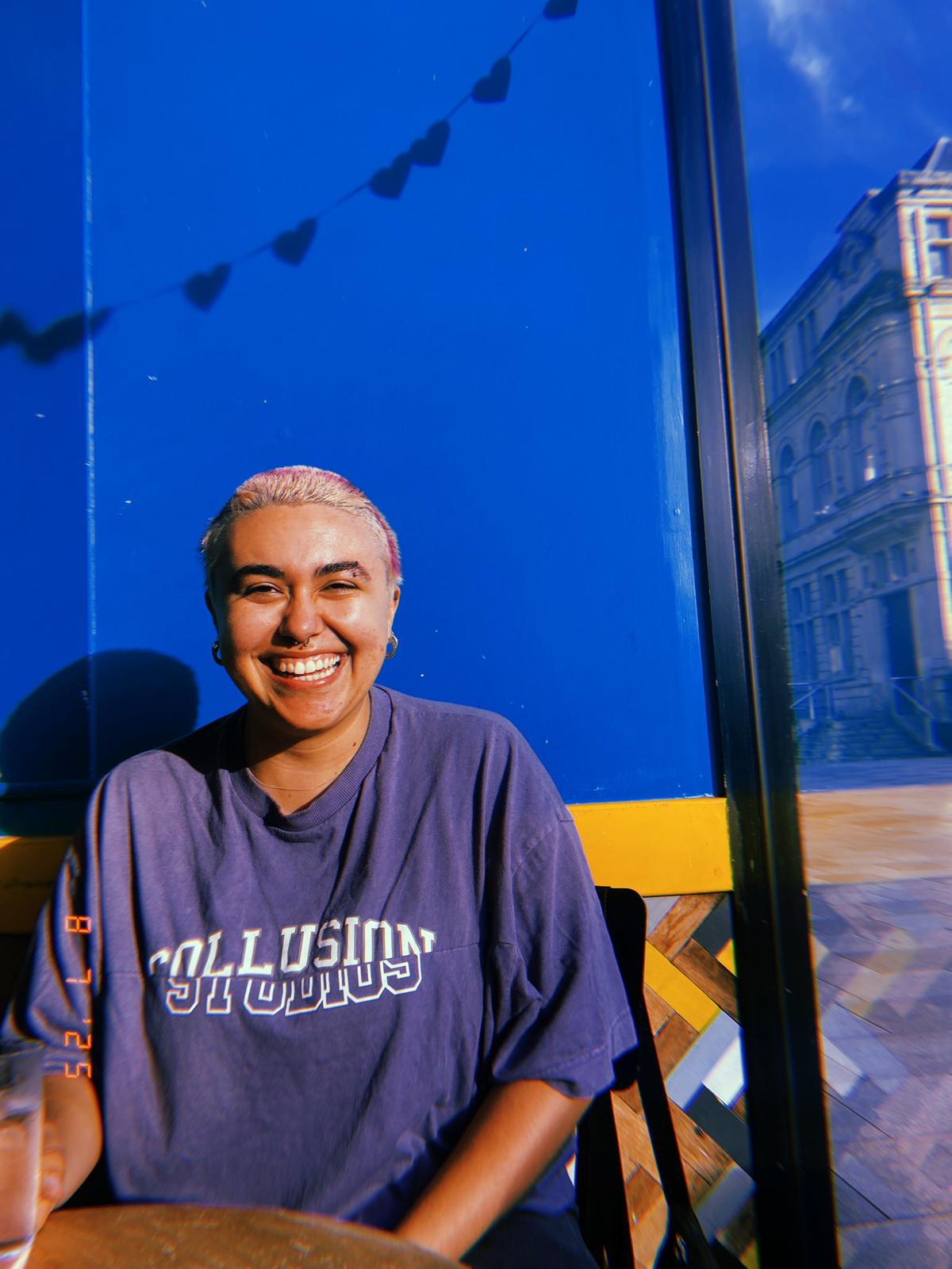 Eugenia is smiling and facing the camera. They are a mixed race person wearing a oversized casual t-shirt and they have short blonde hair. They look friendly and relaxed.