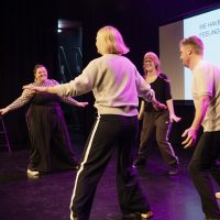 Members of the Sounding Board and Common/Wealth friends take part in a theatre exercise with Gob Squad. They look joyful and appear to be moving in a circle, facing inwards to look at each other.