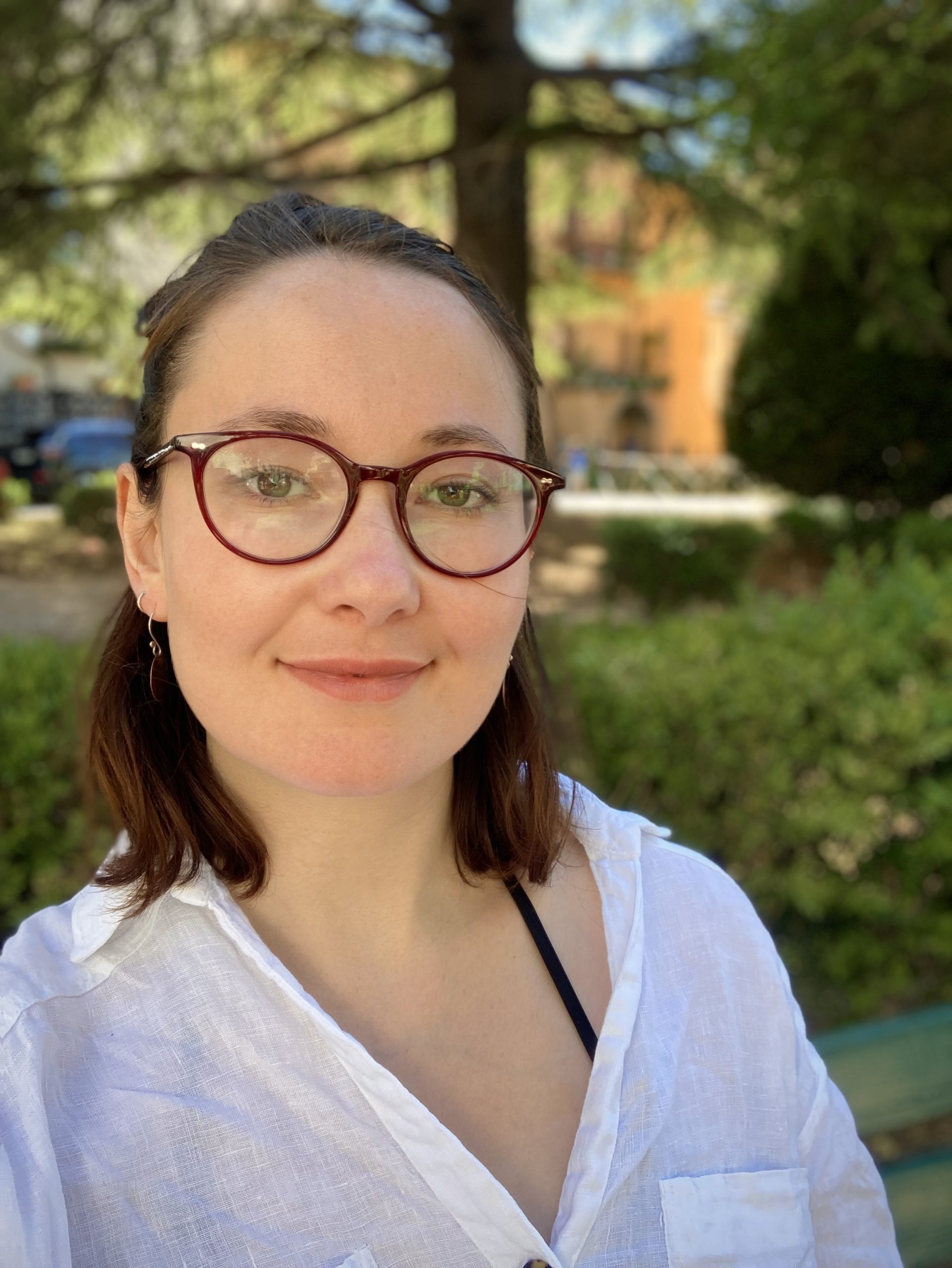 A headshot of a brunette woman wearing red glasses and a white shirt, with trees in the background.